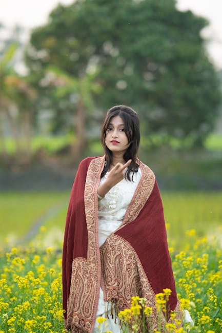 A young woman wearing traditional dress standing amidst vibrant yellow flowers in a lush outdoor setting.