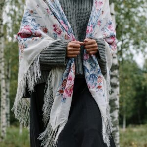 A woman wearing a gray sweater and floral shawl stands amidst serene birch trees, showcasing a stylish look.