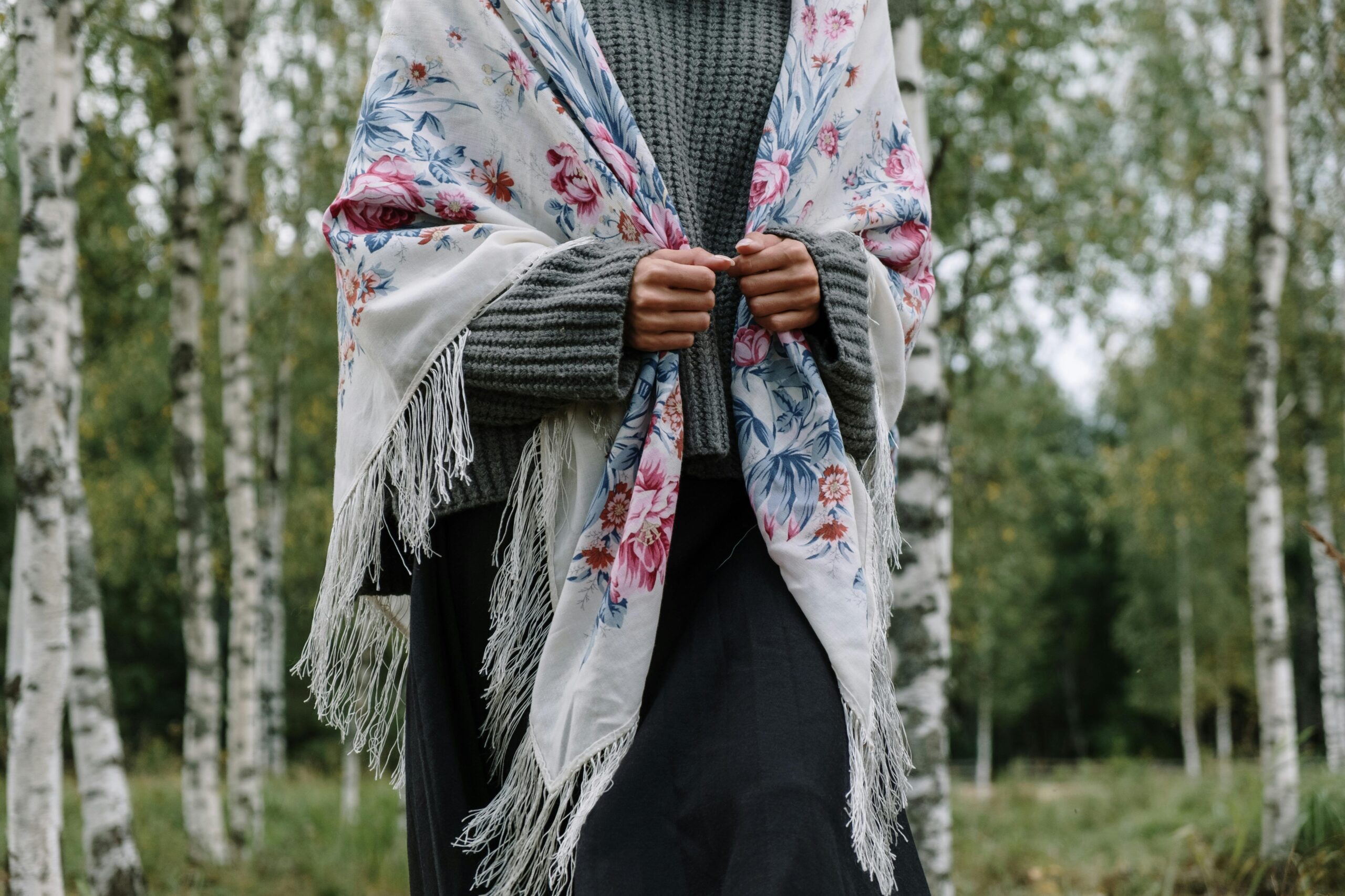 A woman wearing a gray sweater and floral shawl stands amidst serene birch trees, showcasing a stylish look.